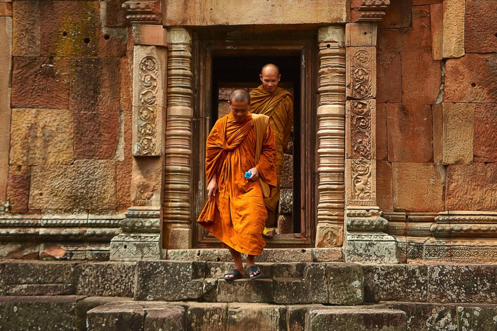 Buddhist monks in orange robes step out of an ancient stone temple in Thailand, showcasing historical architecture.